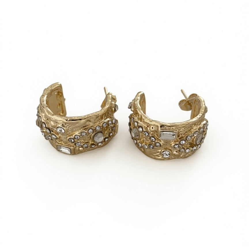 A pair of gold-toned huggie hoop earrings embellished with small clear rhinestones and white stones, shown on a plain white background.